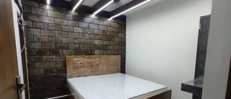 A small, compact room with a single bed, a brick-patterned wall, and a ceiling with exposed beams and lighting.