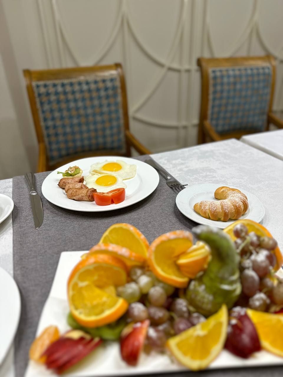 Ein Frühstückstisch mit Tellern voller Speisen, darunter Eier, Fleisch und ein Croissant, sowie eine Obstplatte mit Orangen und Trauben.