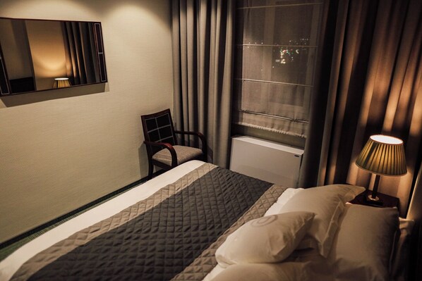 Une chambre d’hôtel comprenant un lit, une chaise, une table de chevet avec une lampe, une fenêtre avec des rideaux et un miroir fixé au mur.