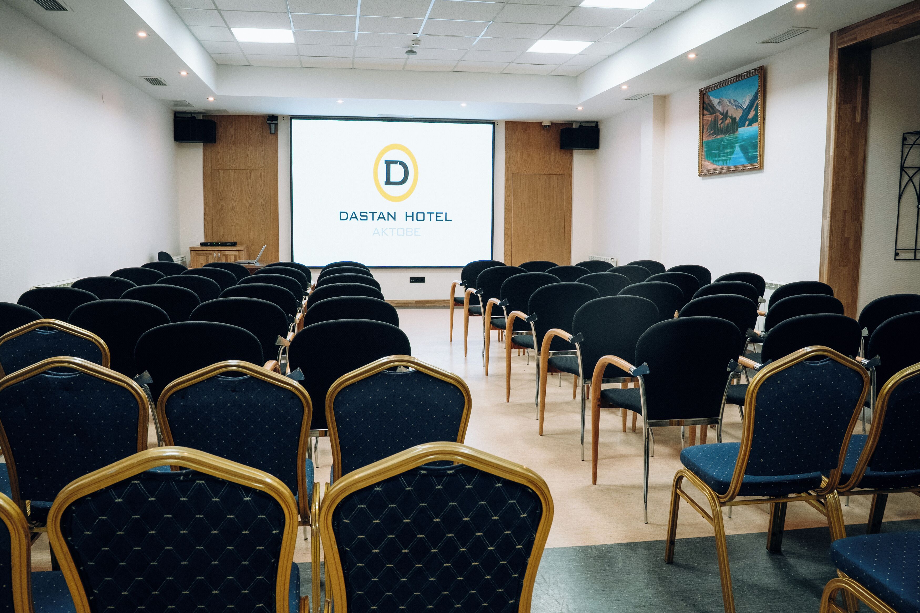 A conference room with rows of chairs facing a projection screen displaying the logo and name of Dastan Hotel.