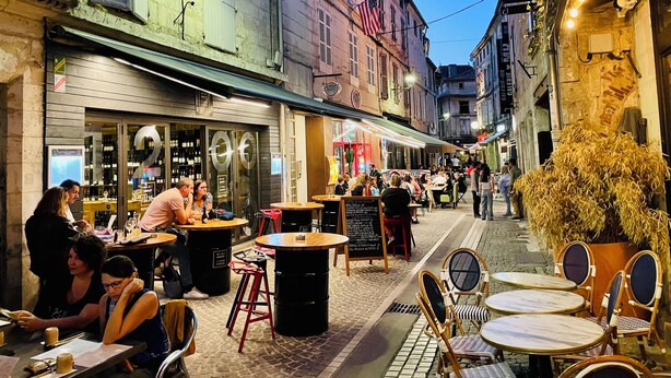 Café al aire libre con mesas y sillas, rodeado de edificios con señalización y un adoquinado.