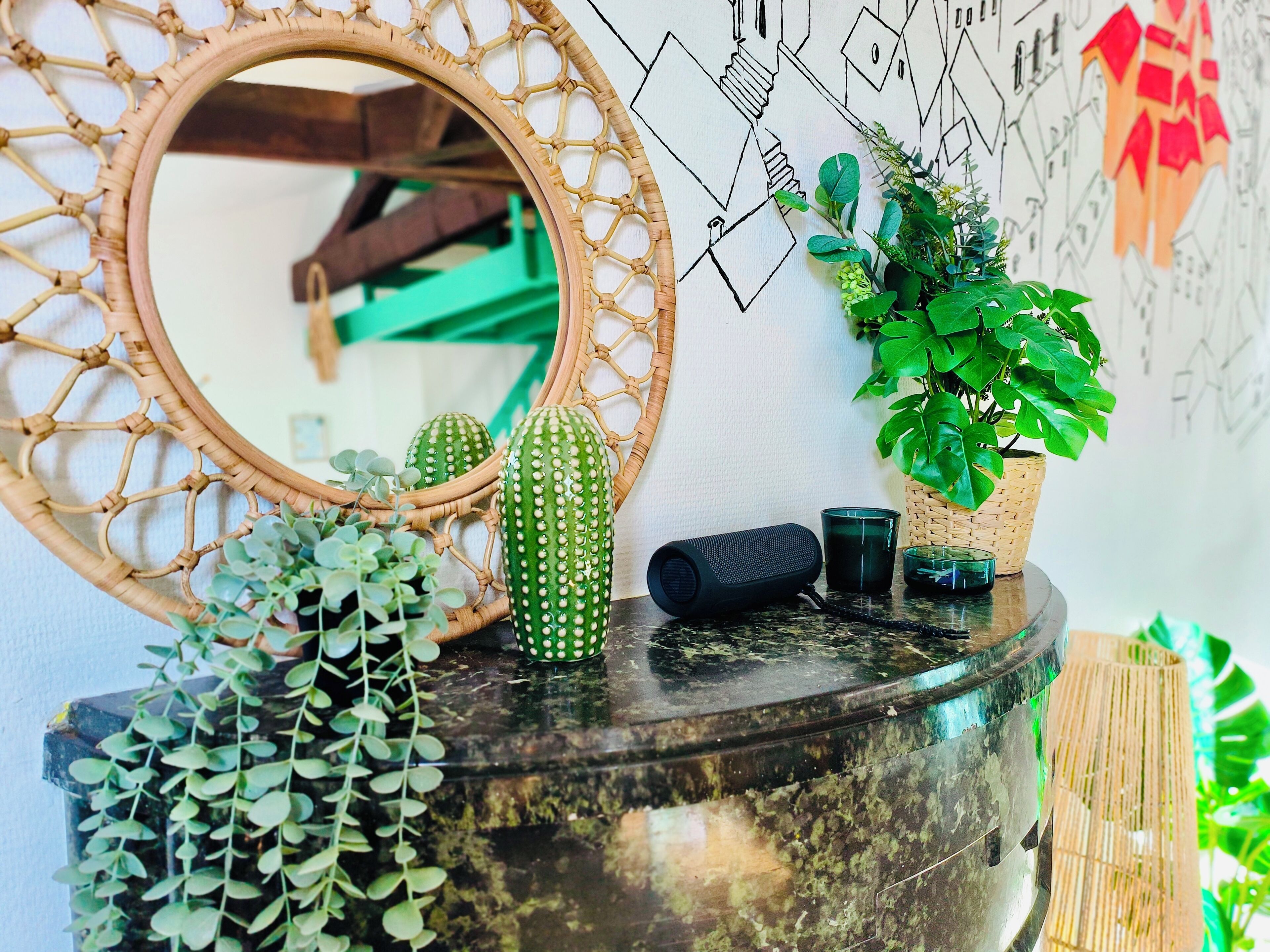 A round mirror with a woven frame, a green potted plant, a black speaker, and a glass cup on a marble surface.