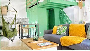 Una sala de estar con una escalera verde, una hamaca colgante, un sofá gris con cojines amarillos y una mesa de centro de madera.