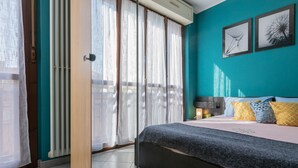 Une chambre à coucher avec un lit, une table de chevet, une fenêtre avec des rideaux et un mur orné de deux tableaux encadrés.