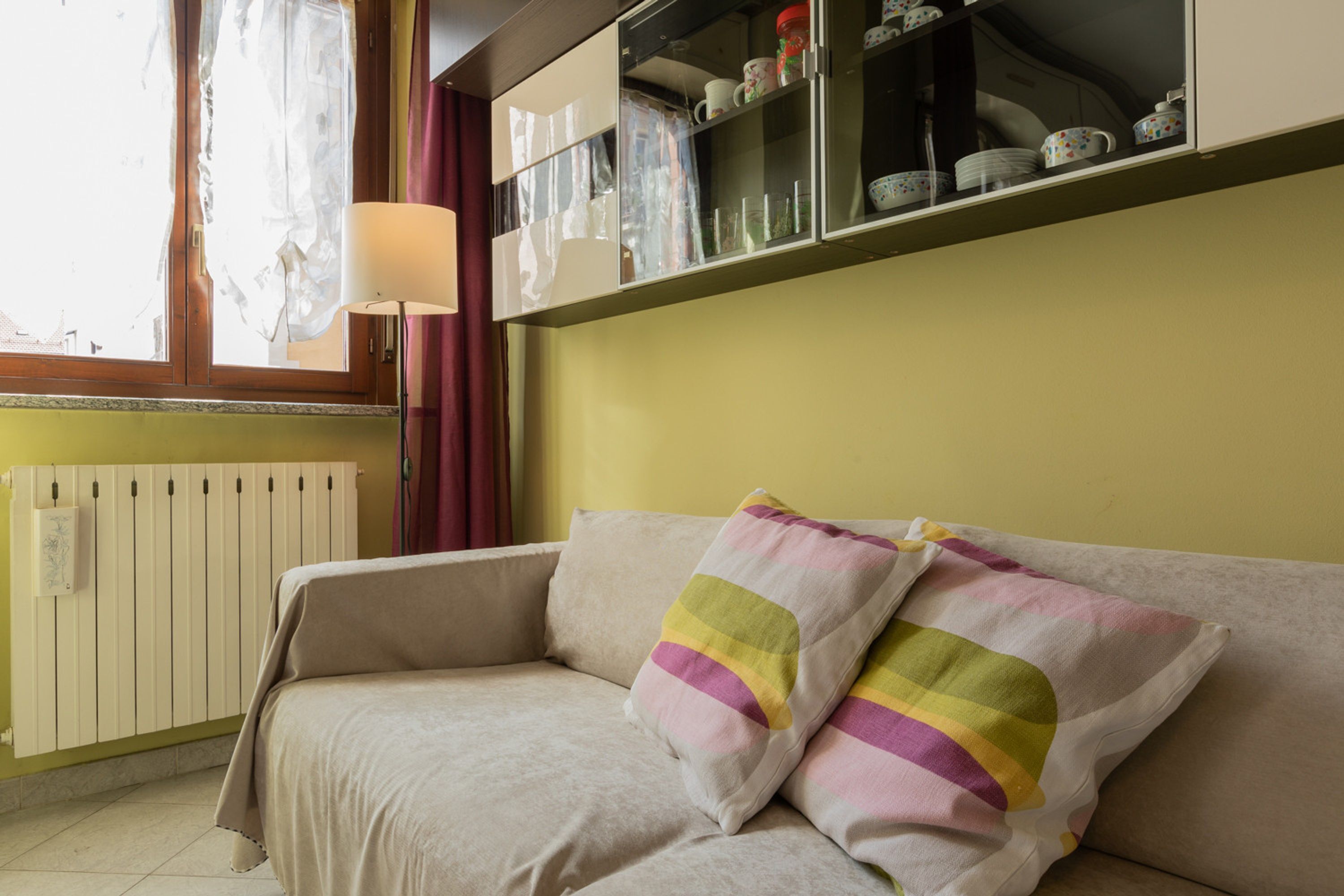 A room with a sofa, a radiator, a window with curtains, and a shelf with dishes.
