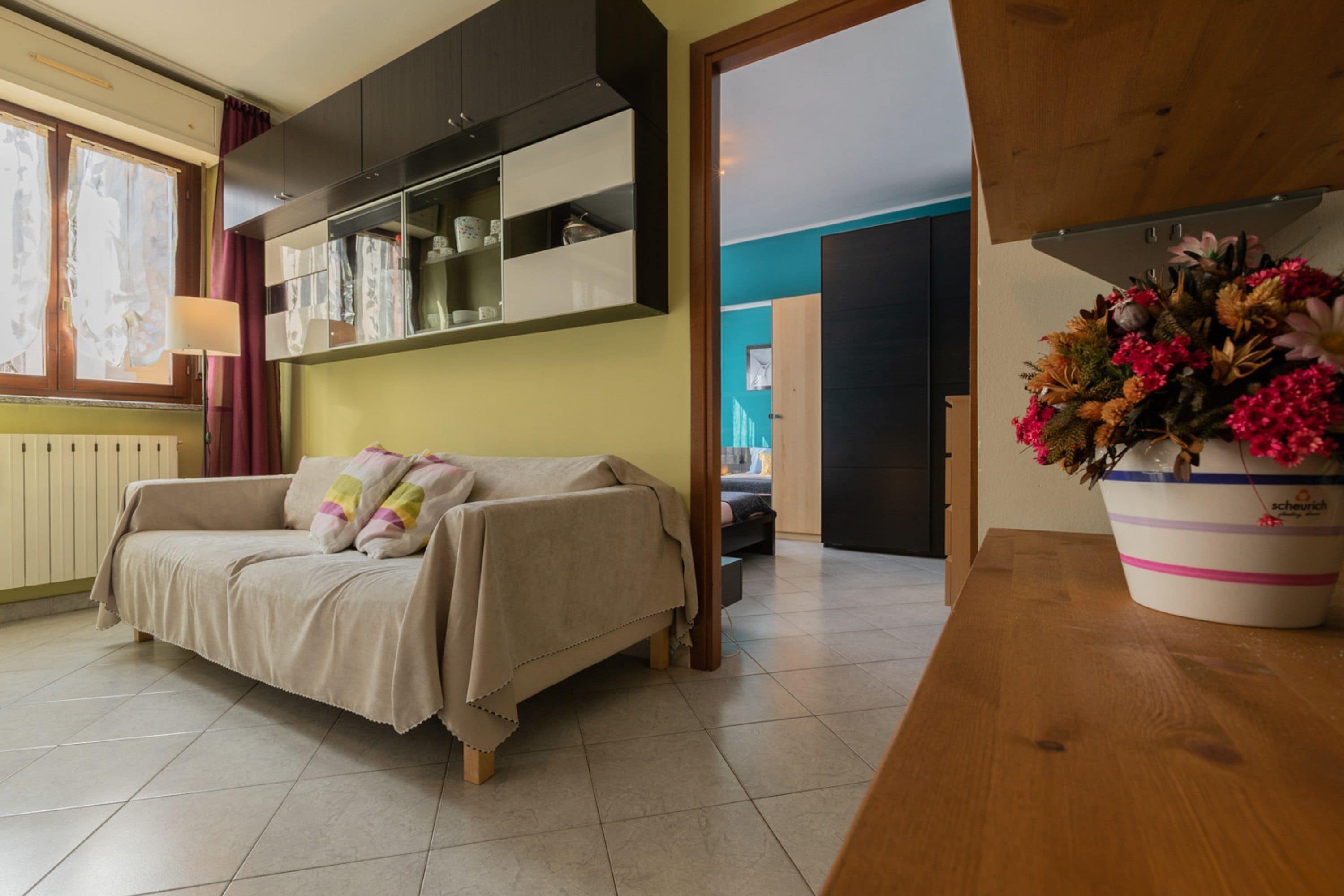 Una sala de estar con un sofá, una mesa de madera, una planta en maceta y un área de cocina con armarios y una ventana.