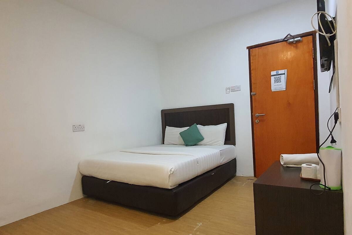 A small, neatly arranged bedroom with a single bed, a wooden nightstand, a door with a QR code, and a power outlet on the wall.