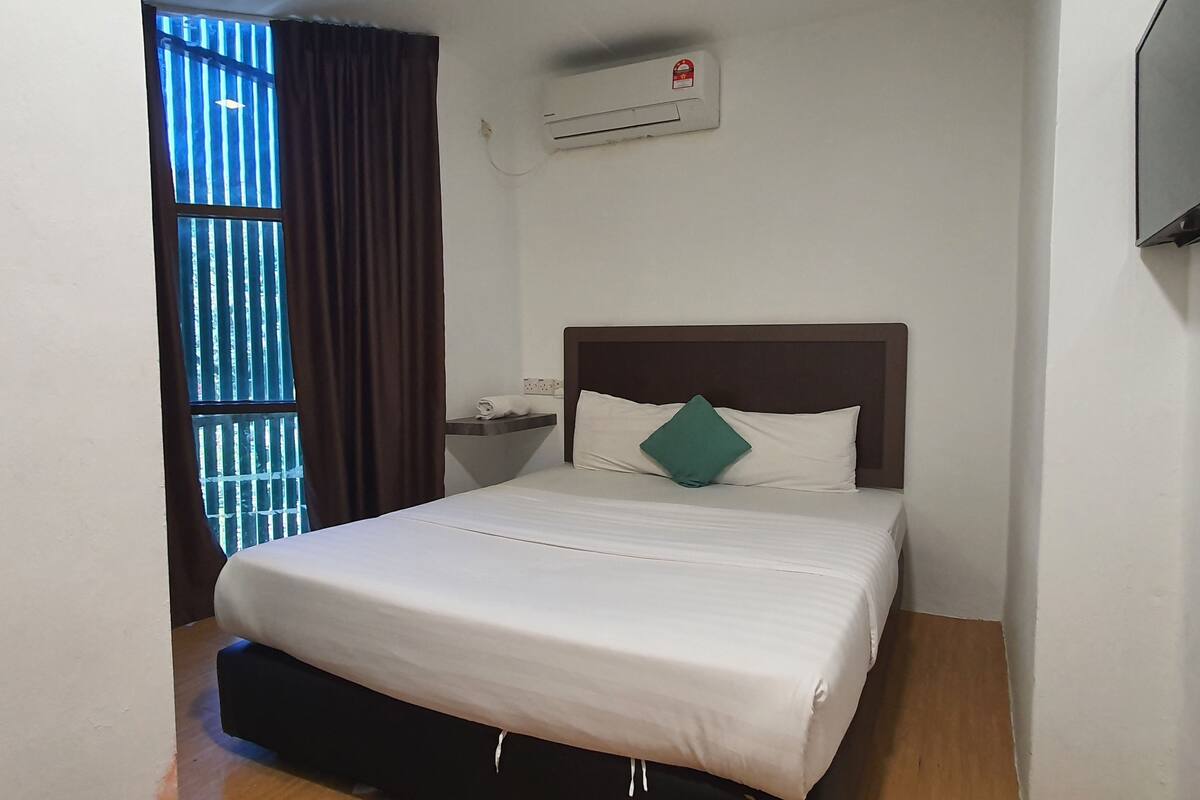 A neatly made bed with a white comforter and a teal pillow, a small bedside table with a lamp, and an air conditioning unit on the wall.