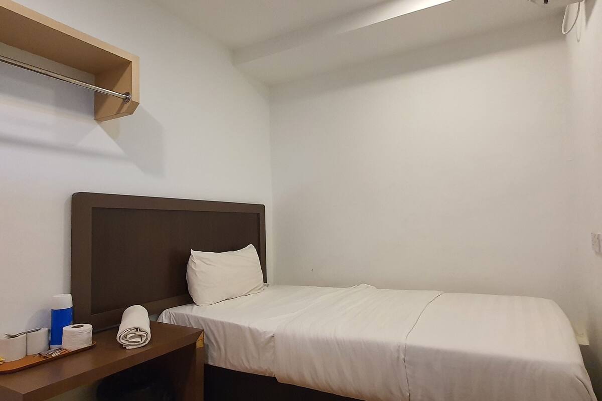 A small, neatly made bed with a wooden headboard, a bedside table with toiletries, and a shelf above the bed.