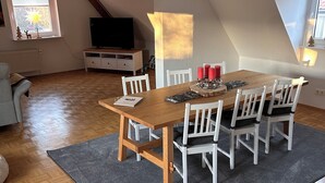 Un comedor con una mesa de madera y sillas blancas, un tapete gris y un televisor al fondo.