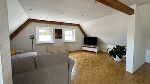 Una habitación amplia en el ático, con techo inclinado, vigas de madera, un ventanal grande, un radiador, un televisor de pantalla plana sobre un soporte blanco, un sofá gris y una planta en maceta.