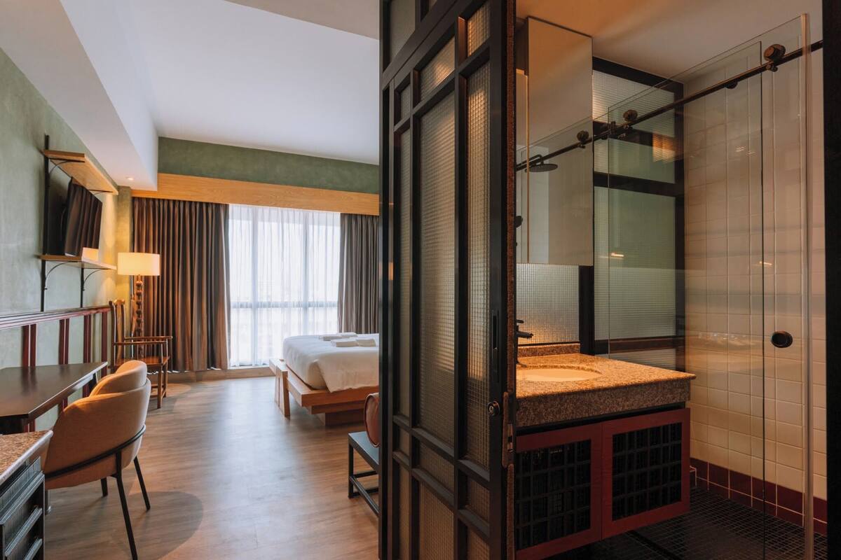 Une chambre d’hôtel comprenant un lit, un bureau, une chaise, une salle de bain avec une douche en verre et une grande fenêtre avec des rideaux.