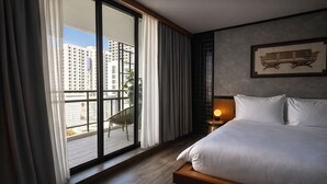 Een hotelkamer met een groot bed, een nachtkastje met een lamp, uitzicht op de stad door een schuifdeur, en een foto in een lijst aan de muur.
