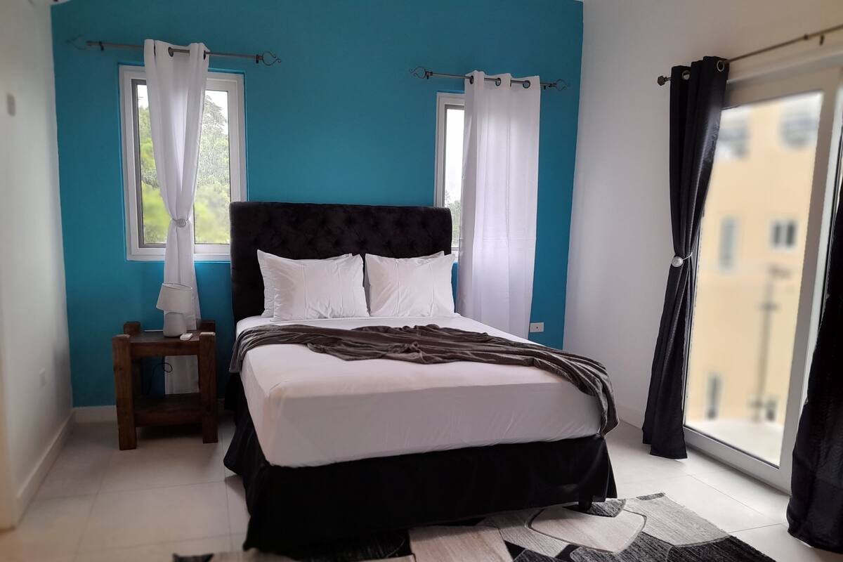 A bedroom with a bed, a nightstand, a window with curtains, and a view of buildings outside.