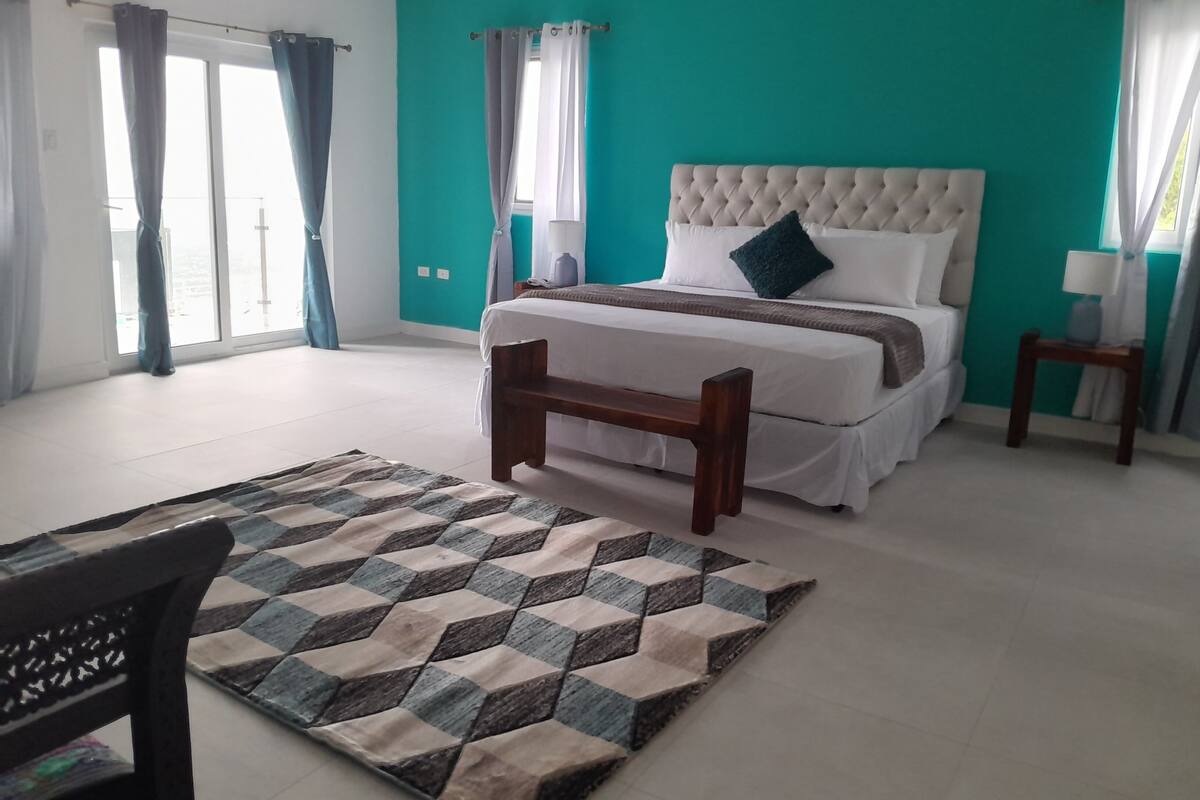 A bedroom with a large bed, a wooden bench, a patterned rug, and a view of the outside through a window.