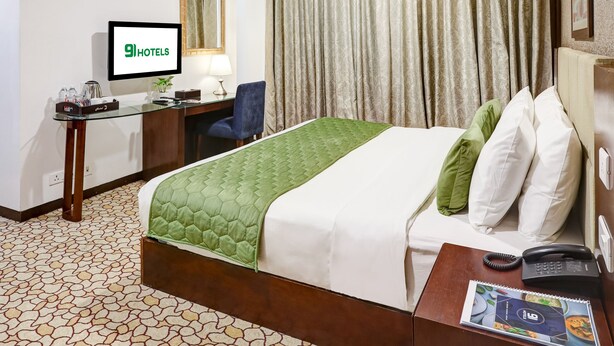 Habitación de hotel con cama, escritorio con computadora, ventilador de techo y televisor mostrando '9HOTELS'.