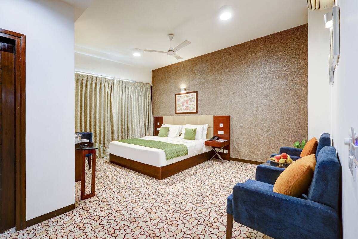 A hotel room with a bed, a blue armchair, a small table, and a ceiling fan.