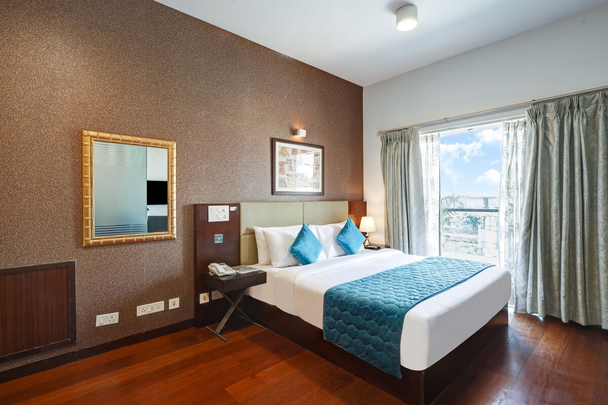 A hotel room with a large bed, a bedside table with a lamp, a mirror, and a window with curtains.