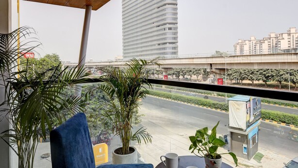 A balcony with a view of a cityscape, featuring a tall building, a bridge, and residential buildings. There are two potted plants and a table with two cups and a small plant. - Hotel 91 Golf Course Road (Gurugram)