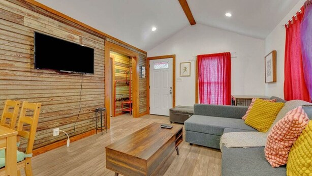 A living room with a wooden wall, a grey sofa, a wooden coffee table, and a television mounted on the wall. - Pink Bungalow! (Austin)