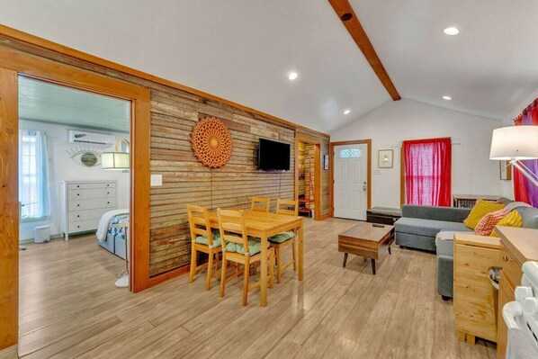A spacious living area with a wooden dining table and chairs, a sofa, and a television mounted on the wall.