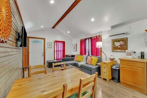 A cozy living room with a wooden dining table, a sofa with colorful cushions, and a television on the wall. - Pink Bungalow! (Austin)