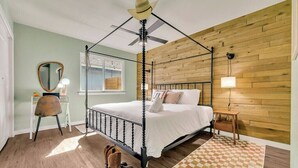 A four-poster bed with a ceiling fan, a wooden wall, a small table with a lamp, and a chair.