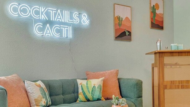 A living room with a neon sign reading 'COCKTAILS & CACTI', a green sofa with cushions, a white coffee table with a cactus, and a checkered floor.