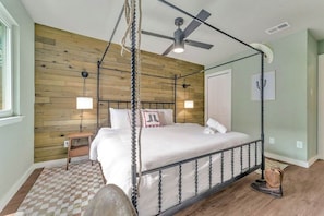A four-poster bed with a ceiling fan, a wooden wall panel, a checkered rug, and a pair of boots on the floor.