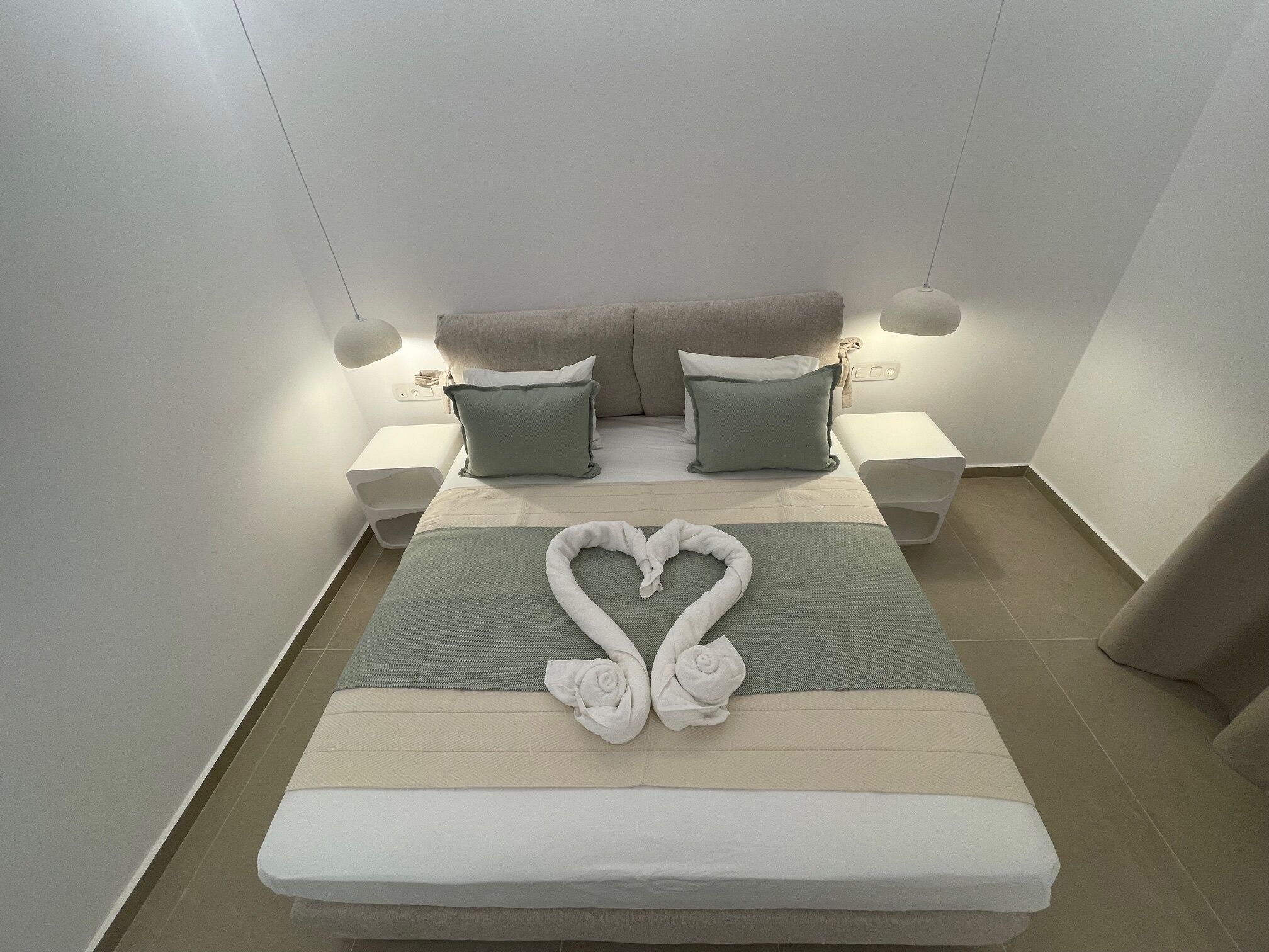 A neatly made bed with a heart-shaped towel arrangement, two bedside tables with lamps, and a neutral color scheme.