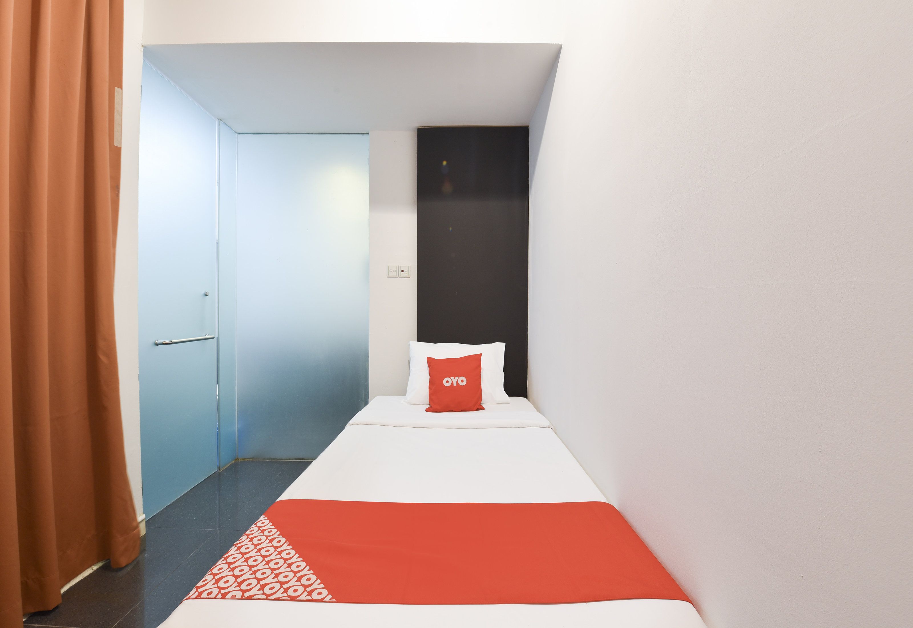 A narrow hotel room with a single bed, a red pillow, and a white blanket.