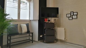 A living room with a black TV stand, a bench with cushions, a potted plant, and a window with a view of buildings. - Laidatxu Beach Apartment in Mundaka (Mundaka)