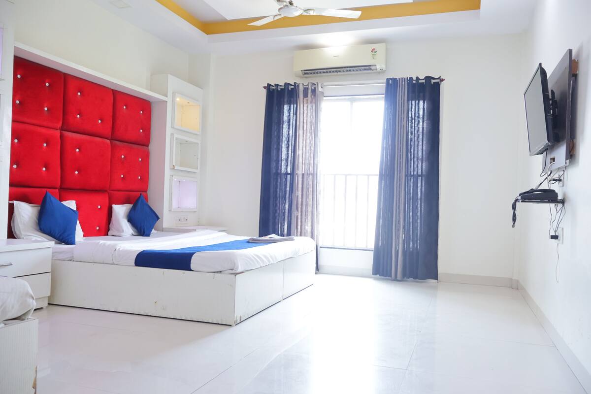 A hotel room with a large bed, a ceiling fan, an air conditioning unit, a TV mounted on the wall, and a window with curtains.