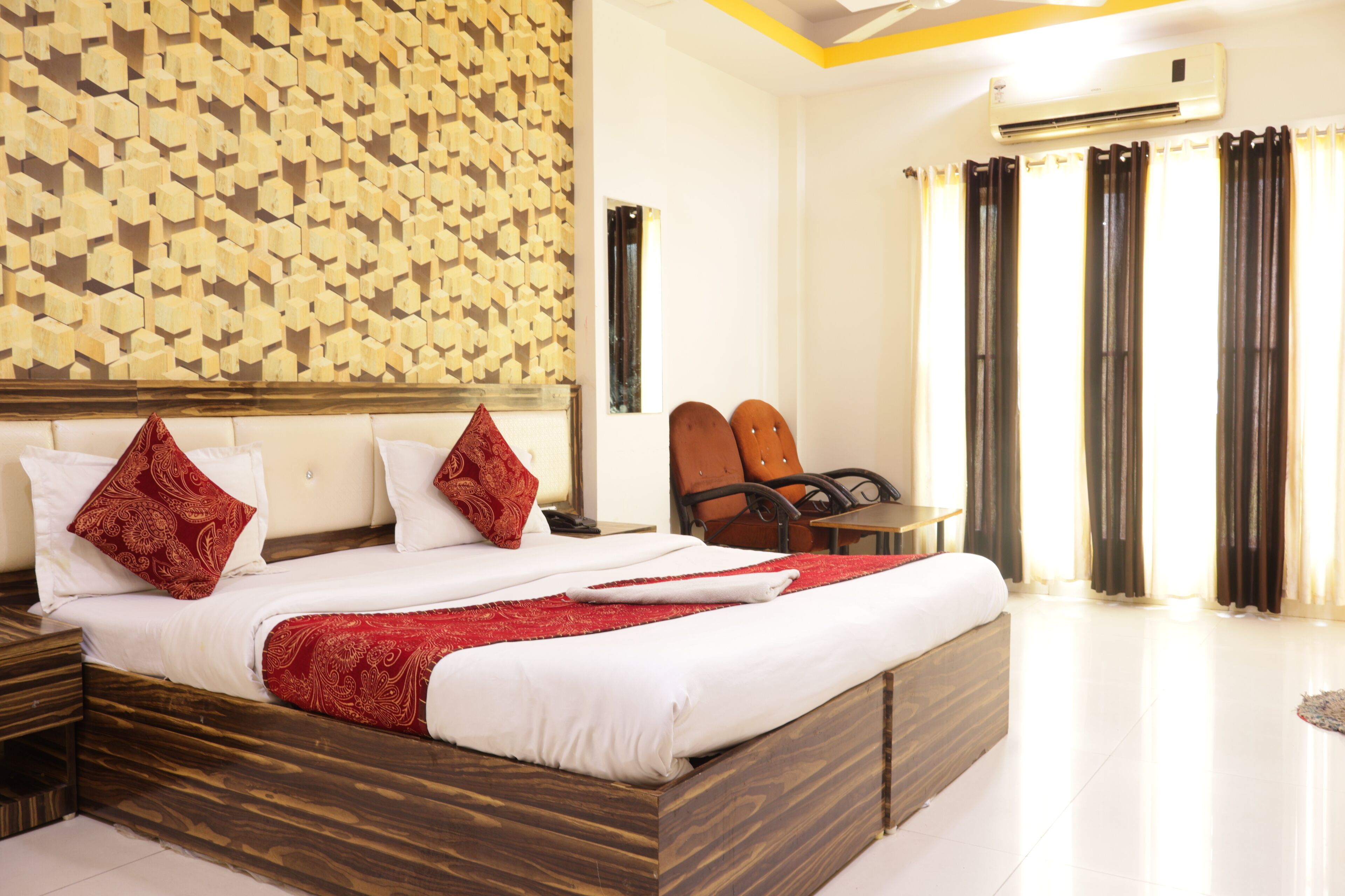 A hotel room with a large bed, a wooden headboard, a patterned wall, a chair, a small table, and a window with curtains.