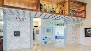 A kitchen with a herringbone tile backsplash, a built-in dishwasher, and a wooden shelf above the countertop holding various bottles and glasses.