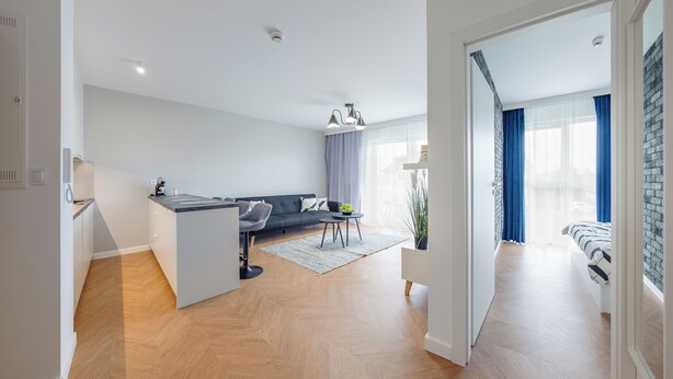 A modern apartment with a kitchen, dining area, and living room. It features a herringbone wood floor, a grey sofa, and a dining table with chairs. - Apartamenty Sun & Snow Kuracyjna Jastrzębia Góra (Wladyslawowo)
