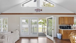 A spacious living area with a high ceiling, a sliding glass door leading to an outdoor deck, a wooden floor, and a kitchenette with wooden cabinets. - Serenity on the Lake (Prosperity)