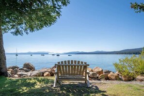 A wooden bench by a lake with rocks and a tree, boats in the water, and distant mountains.