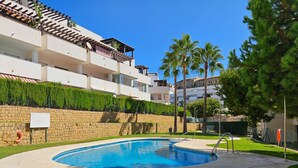 A swimming pool with a stone wall and greenery, surrounded by a building with balconies and a clear blue sky. - Two bedroom apartment in Riviera del sol (Mijas Costa)