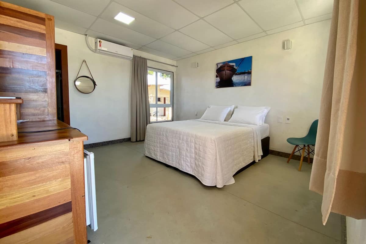 Habitación de hotel con cama, una silla, un escritorio de madera, equipo de aire acondicionado y ventana con cortinas.