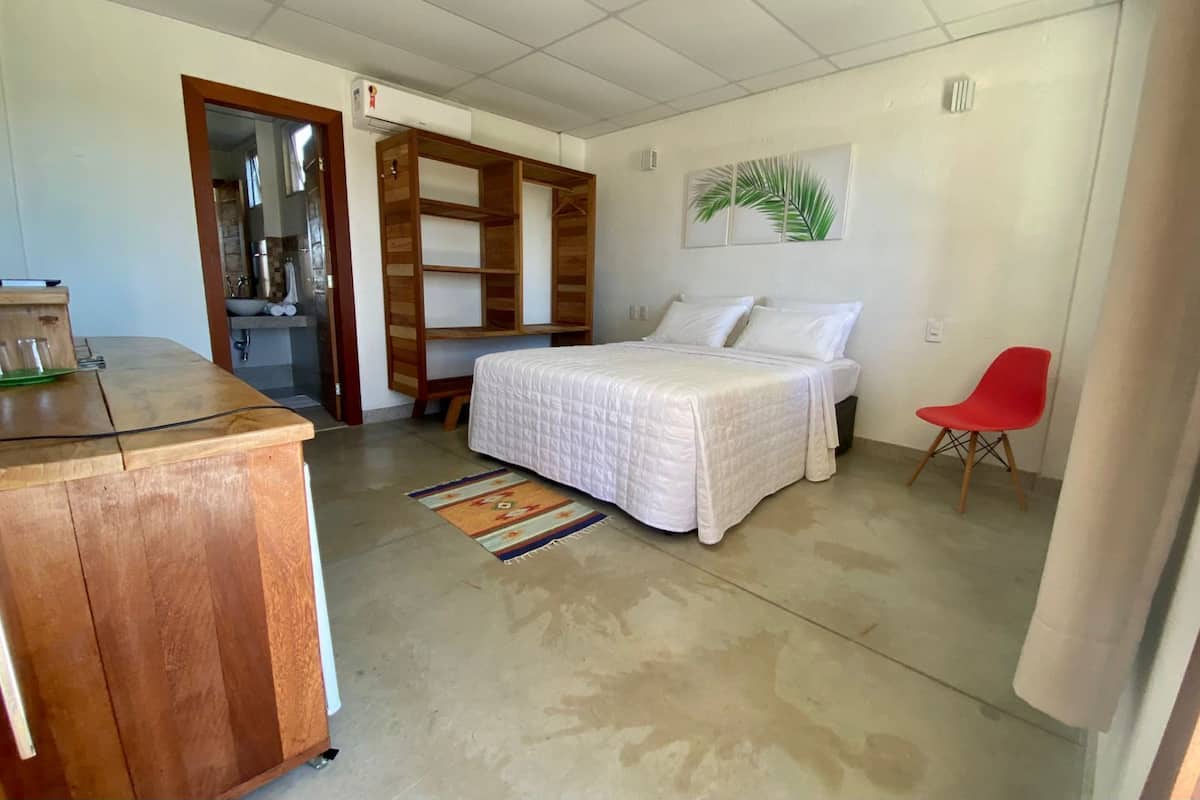 Un dormitorio con una cama, una silla roja, un escritorio de madera y un baño visible a través de una puerta abierta.