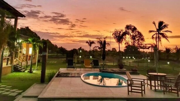 A pool area with lounge chairs and a small building, surrounded by palm trees and a sunset sky. - Camaleao Ecopousada Ltda (Linhares)