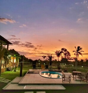 A pool area with lounge chairs and a small building, surrounded by palm trees and a sunset sky.