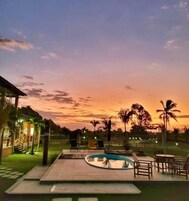 Área de piscina com espreguiçadeiras e um pequeno edifício, cercada por palmeiras e um céu ao pôr do sol.