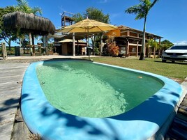 Uma piscina com deck de madeira, uma estrutura de palha e um edifício com guarda-sol amarelo ao fundo.
