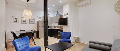 A modern living room with a sofa, a coffee table, and two blue chairs. There is a kitchen area with a dining table and chairs, a TV, and a small side table.