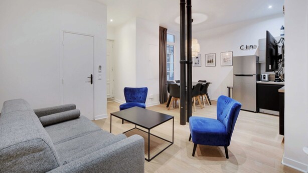 A modern living room with grey sofas, a black coffee table, and blue chairs. There is a dining area with a table and chairs, and a kitchen with a refrigerator and a TV. - Elegant Apartment - 2bd/6p - Hotel de Ville (Paris)