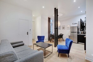 A modern living room with grey sofas, a black coffee table, and blue chairs. There is a dining area with a table and chairs, and a kitchen with a refrigerator and a TV.