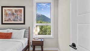Une chambre à coucher comprenant un lit, une table de chevet, une lampe, un tableau encadré et une fenêtre donnant sur une vue sur les montagnes.