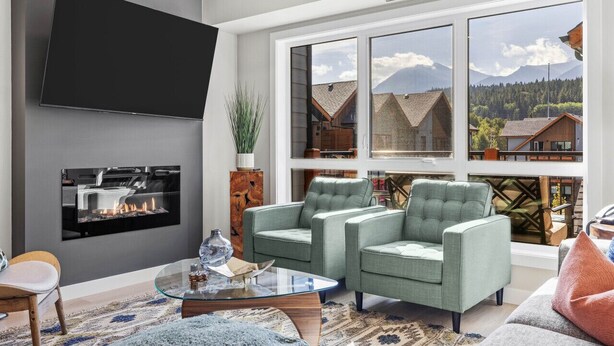 Un salon moderne doté d’un téléviseur à écran plat, d’une cheminée, de deux fauteuils verts, d’une table basse en verre et de grandes fenêtres offrant une vue sur les montagnes.